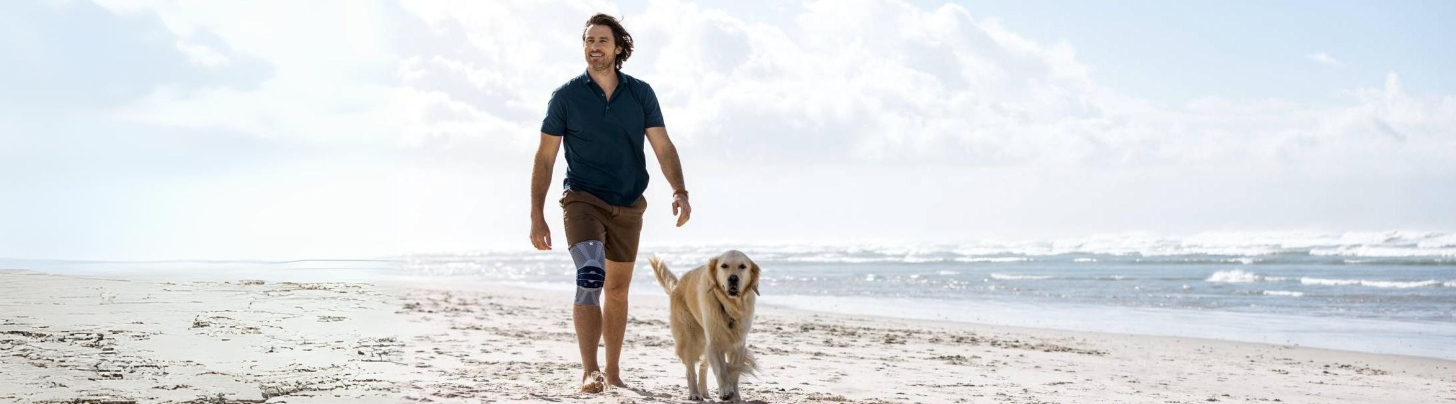 Man sitting with his dog on the beach. He is wearing Bauerfeind's GenuTrain Knee Brace, a good fit for supporting the knee and minimising knee clicking