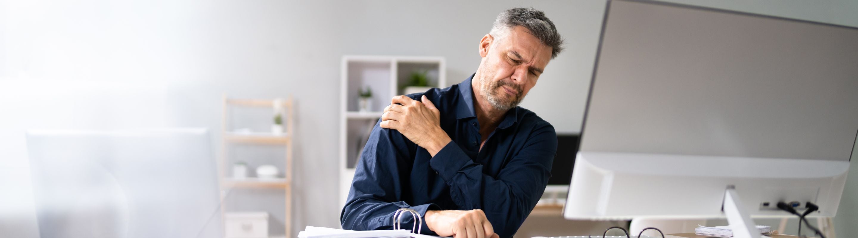Man holding his shoulder in pain. He is seated at a work desk