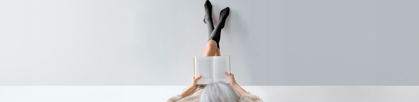 woman wearing compression socks propping her feet up against a wall while reading a book