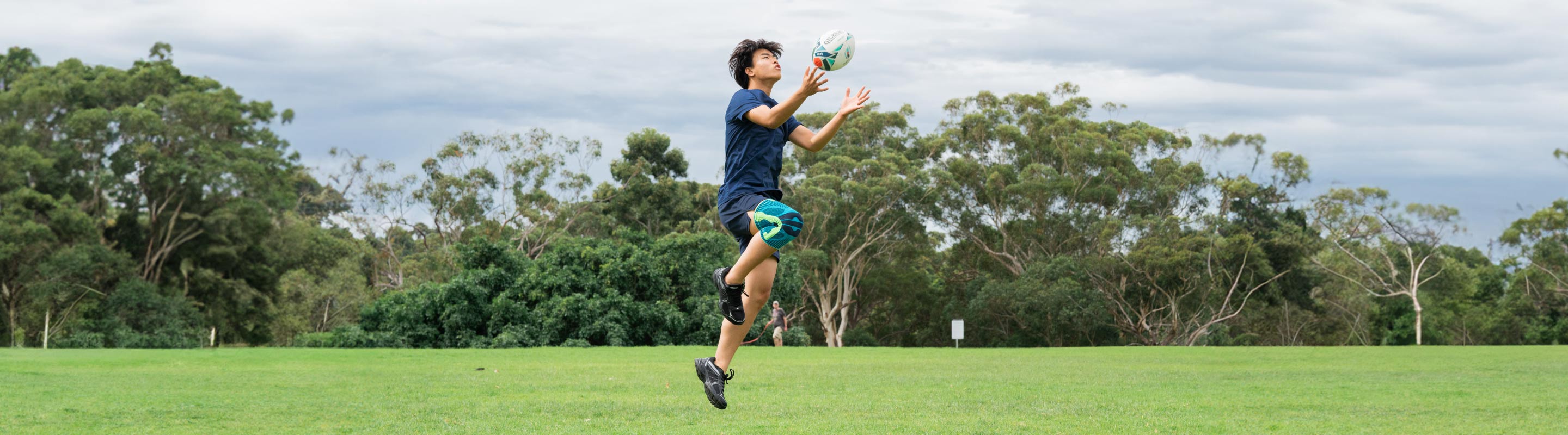 Young man playing ruby at the park. He is jumping up and catching a rugby ball while wearing Bauerfeind's Sports Knee Support