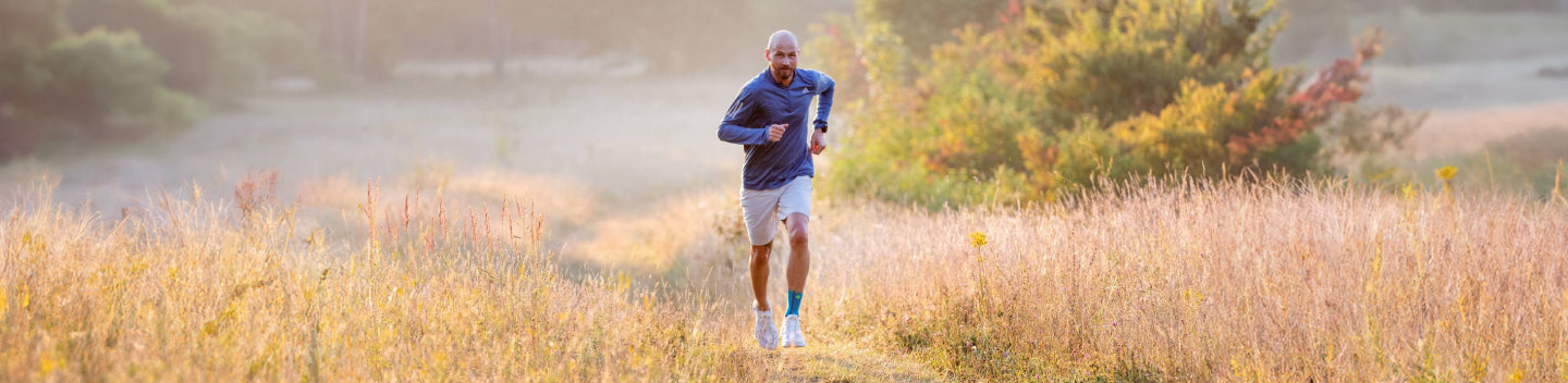 Man jogging through a grassy field. He's wearing a blue long sleeve shirt, white running shoes, a Bauerfeind sports ankle brace