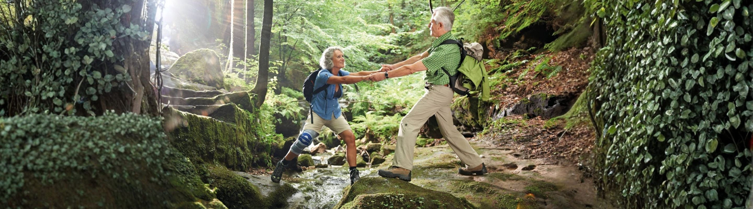 man and woman hiking. The man is helping the woman over a small river. The woman is wearing Bauerfeind's GenuTrain S Knee Brace