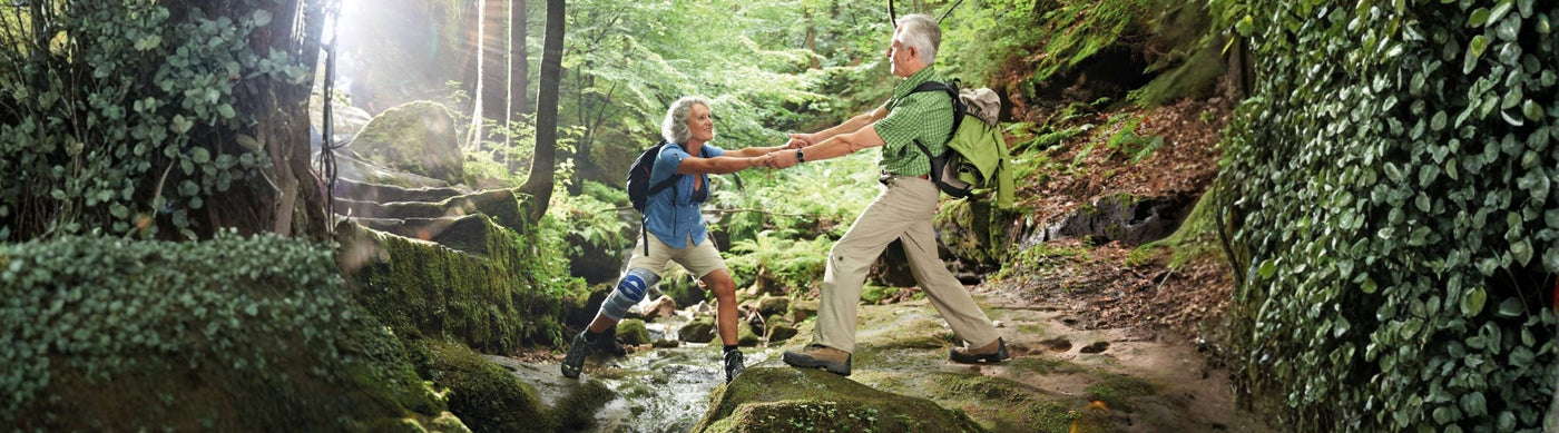 man and woman hiking. The man is helping the woman over a small river. The woman is wearing Bauerfeind's GenuTrain S Knee Brace