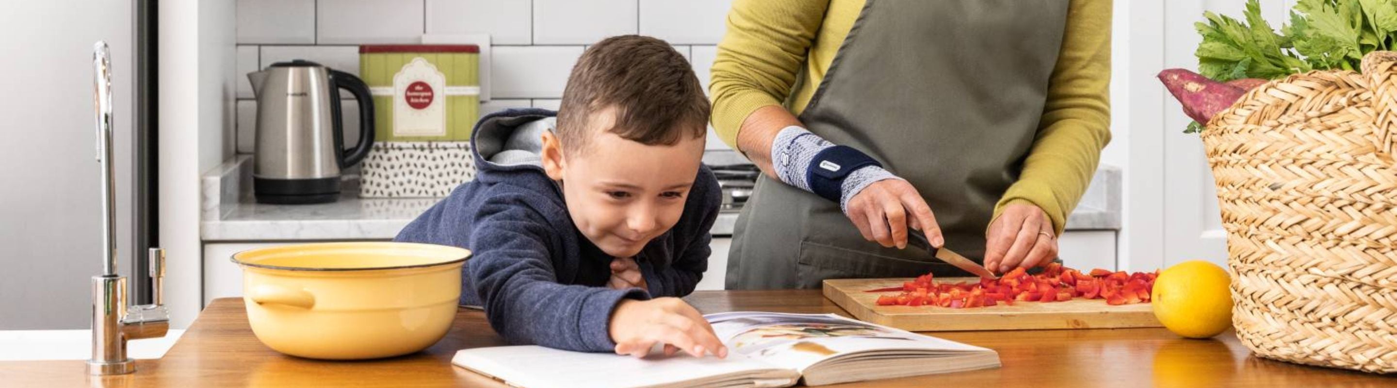 woman chopping vegetables in the kitchen as a child leans over the counter and looks at an open cook book. The woman is wearing a ManuTrain Wrist Brace for carpal tunnel pain