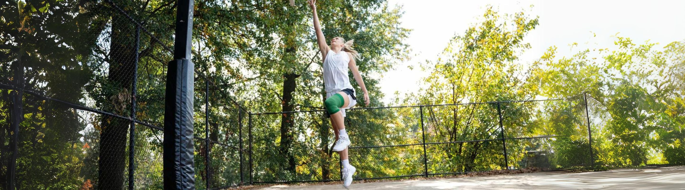 person in the middle of making a shot on an outdoor basketball court. She is wearing Bauerfeind's NBA Compression Knee Support and basketball shoes, essential basketball training gear