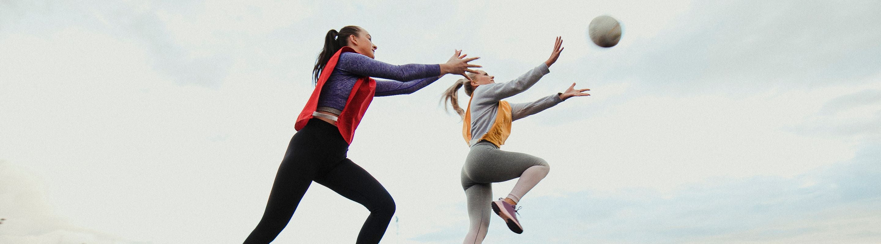 Two girls playing netball. They are on opposite teams, jumping up to catch the ball at the same time. 