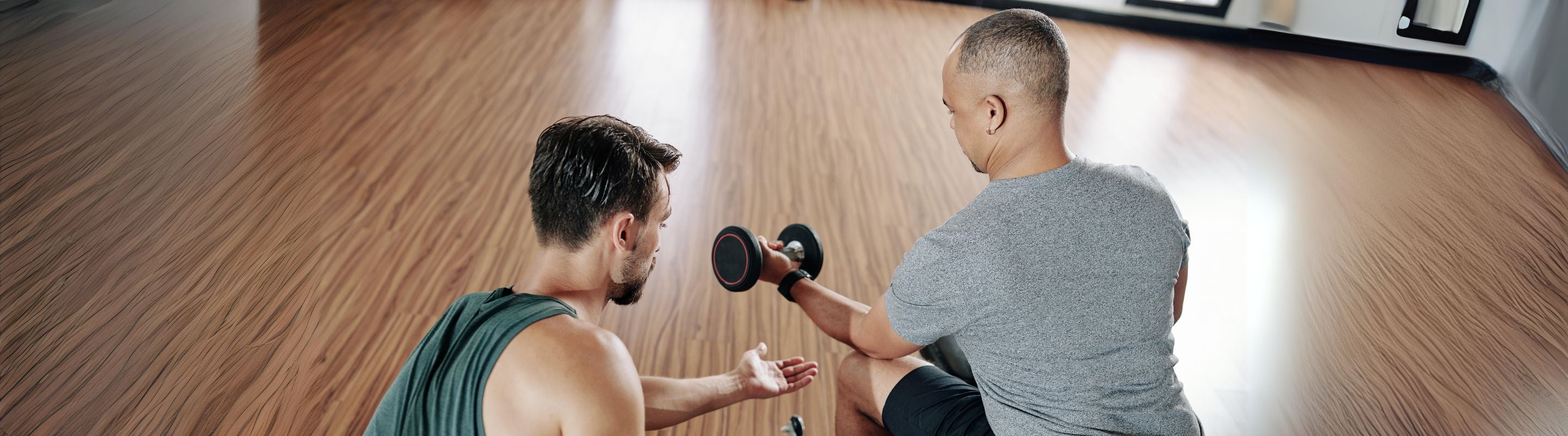 men at the gym. one is showing the other how to do resisted wrist flexion exercises with a dumbell