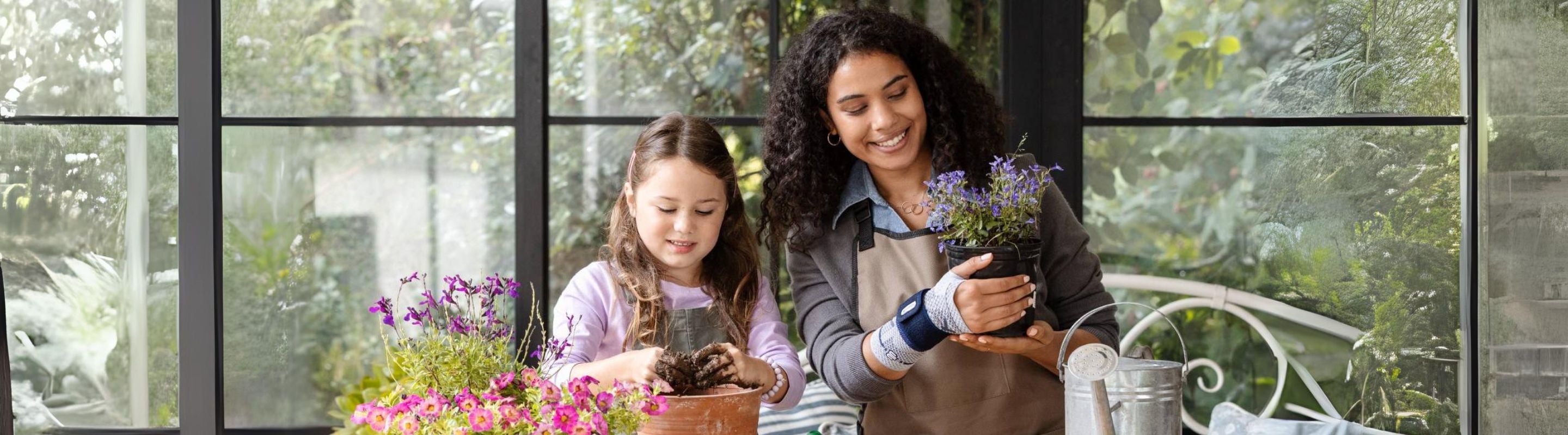 Woman and a girl gardening. Each is smiling and holding a potted plant. The woman is wearing a Bauerfeind ManuTrain Wrist Brace for her repetitive strain injury