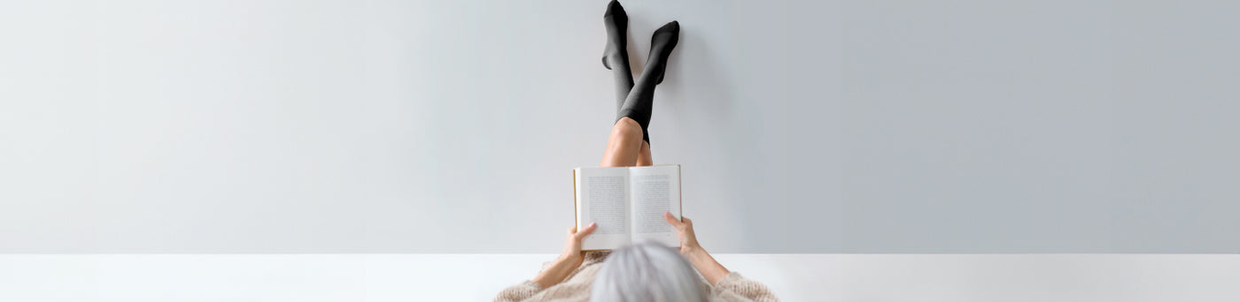 woman wearing compression socks propping her feet up against a wall while reading a book
