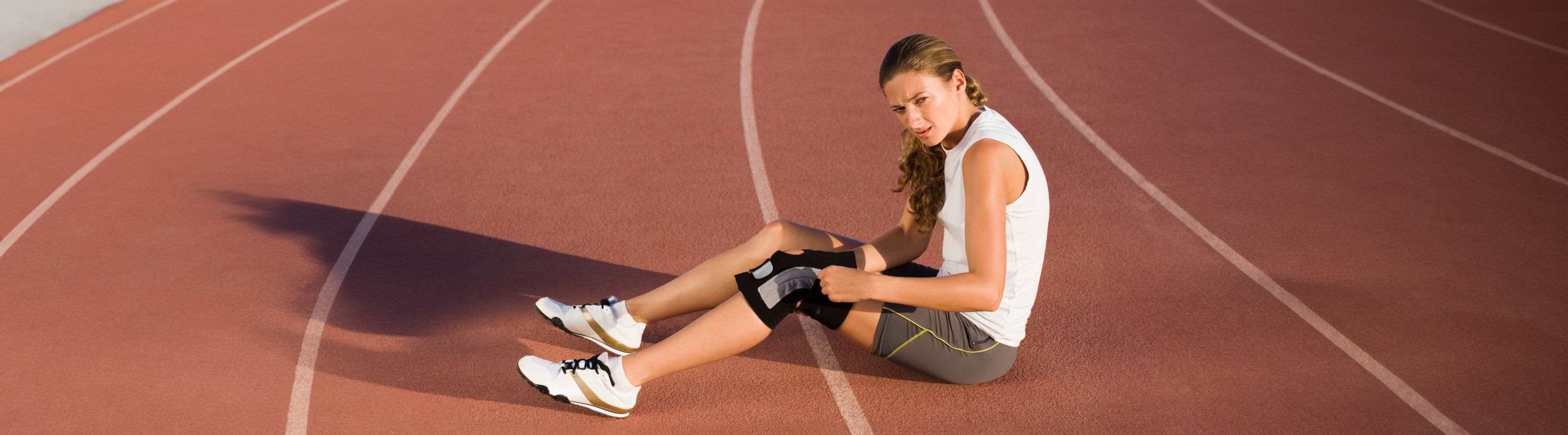 Woman sitting down unhappily on a track field with her legs in front of her, trying to adjust her neoprene knee brace