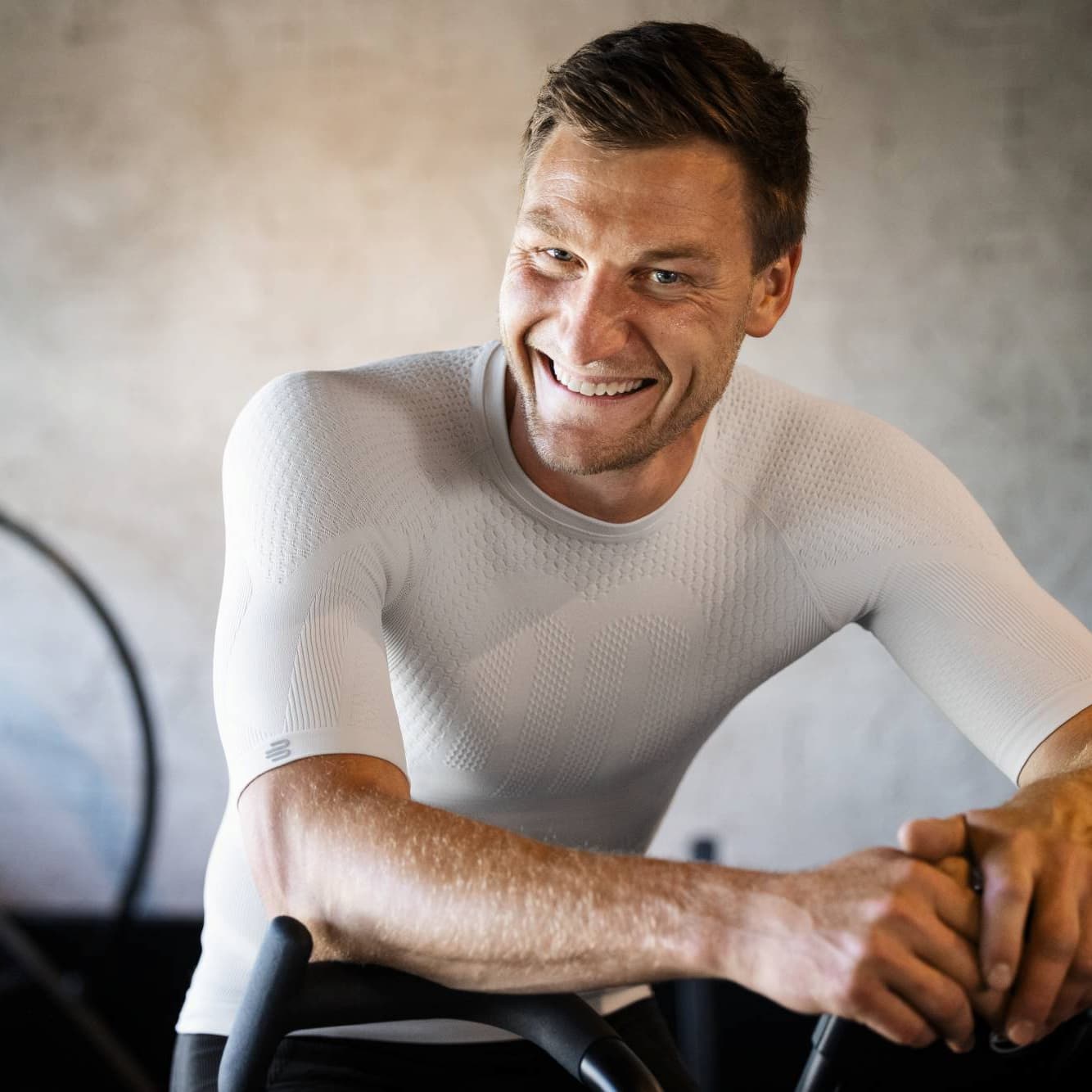 Man wearing a fitted silver Bauerfeind compression shirt sitting on a bike indoors.
