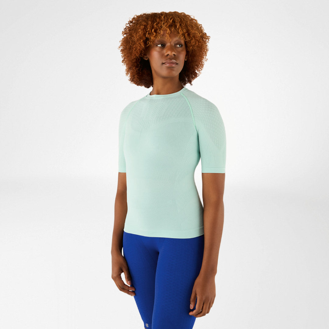 Woman wearing a light green Bauerfeind compression shirt and blue leggings on a white background.